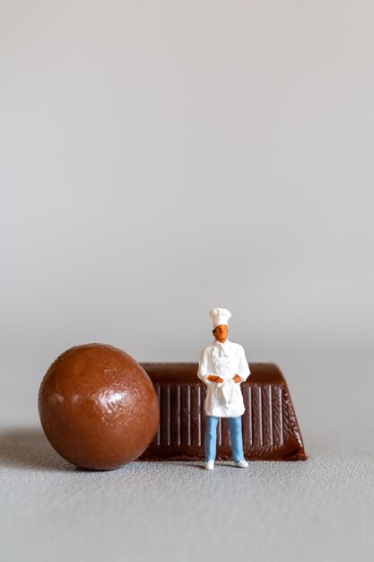 Miniatuurmensenchef-kok met chocolade die zich bevindt terwijl hij tegen een grijze achtergrond staat