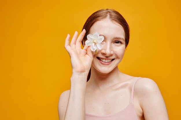 Minima portrait of young red haired woman with flower smiling at camera against vibrant yellow backg