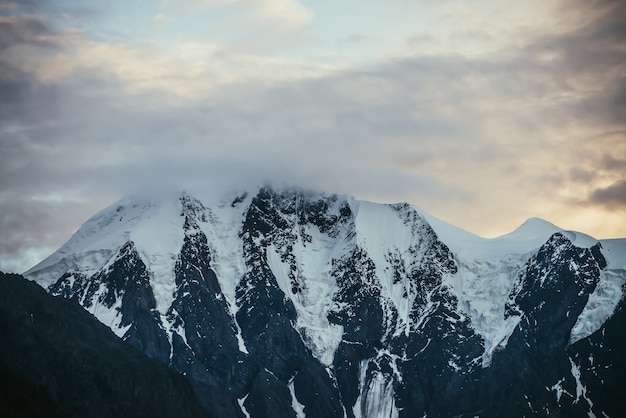 Minimaal alpenlandschap met besneeuwde bergtop onder lage wolken op de achtergrond van zonsondergang of zonsopgang bewolkte hemel van verhelderende kleur. Sfeervol landschap met sneeuwwitte bergrug in de dageraadhemel.