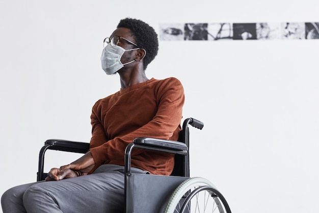 Minimaal portret van een afro-amerikaanse man die een rolstoel gebruikt en een masker draagt terwijl hij naar schilderijen in de galerie voor moderne kunst kijkt,