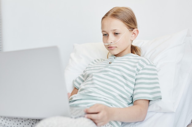 Minimaal portret van een klein meisje dat in het ziekenhuisbed ligt en een laptop gebruikt met zuurstofondersteuning, kopieer ruimte