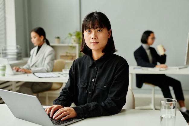 Minimaal portret van jonge Aziatische zakenvrouw die laptop gebruikt en naar de camera kijkt met collega's op de achtergrond