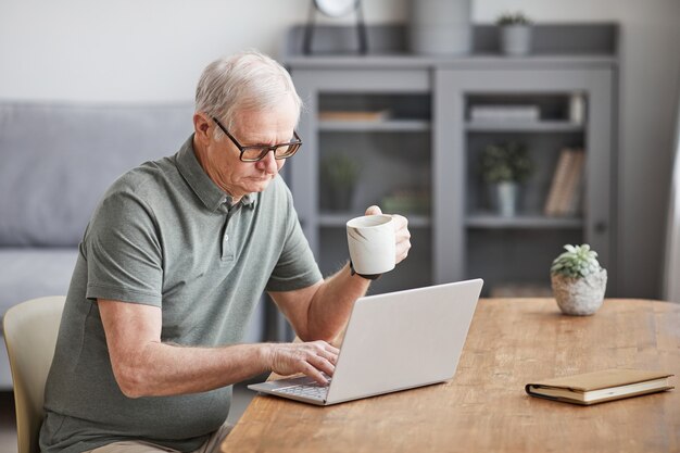Minimaal portret van moderne senior man die thuis laptop gebruikt en geniet van koffie, kopieer ruimte