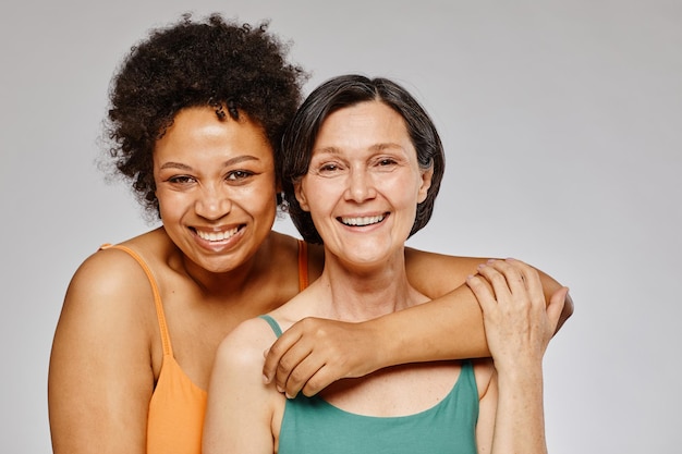 Minimaal taille-up portret van twee echte vrouwen die elkaar omhelzen en vrolijk glimlachen tegen een grijze achtergrond