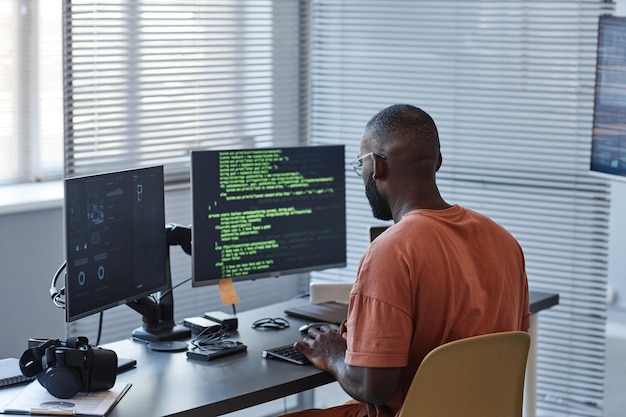 Minimal back view of african american man writing code in green lines and using computers in office