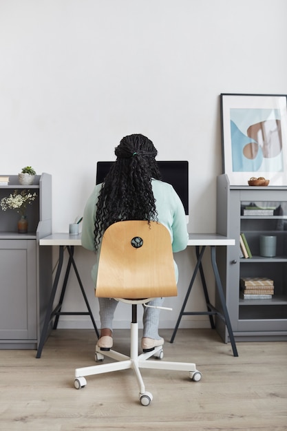 Foto vista posteriore minima di una donna afroamericana formosa che utilizza il pc sul posto di lavoro di casa in interni moderni, spazio di copia