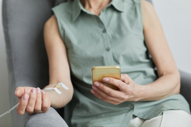 Minimal close up of unrecognizable woman getting IV drip and using smartphone, copy space