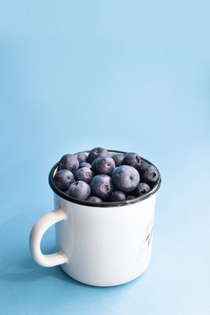 Minimal composition of fresh blueberries in a white metal mug on a blue background.