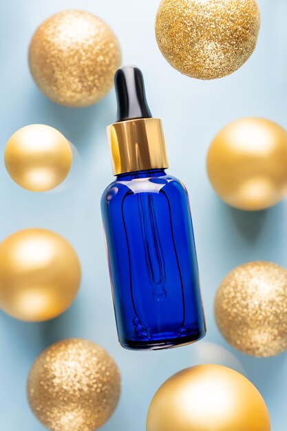 Minimal composition with serum or oil in blue glass bottle with dropper on creative background with golden christmas baubles Winter skin treatment concept