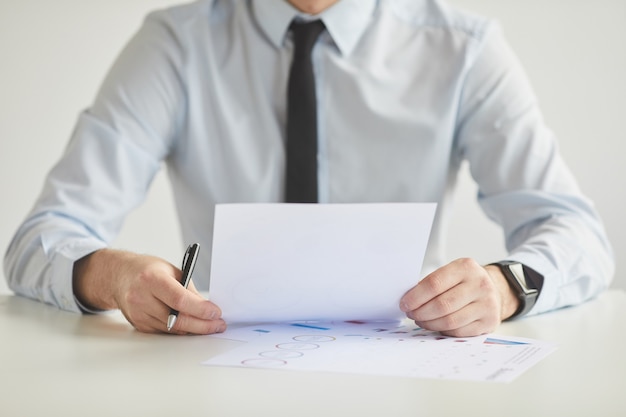 Minimal cropped portrait of unrecognizable young businessman holding document and while working sitting at desk and in office against white wall, copy space