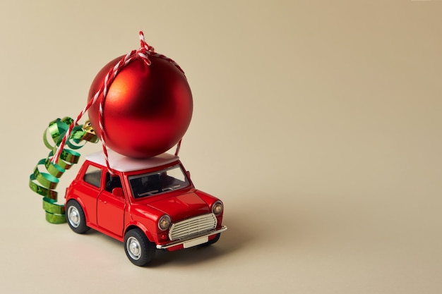 Minimal design for celebrating christmas or new year greeting card. Delivery of decorations and gifts. Little red toy car and Red Christmas ball on a light background