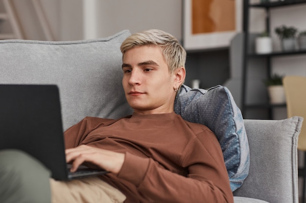 Minimal portrait of blonde young man using laptop while lying on sofa in home interior, copy space