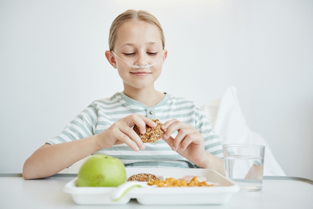 Ritratto minimo di ragazza che mangia un pranzo sano nella stanza d'ospedale bianca e sorride