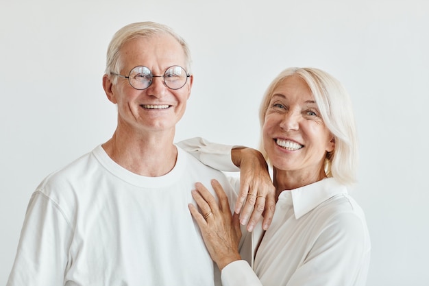 白い背景に白を着て、カメラに微笑んでいる現代の年配のカップルの最小限の肖像画