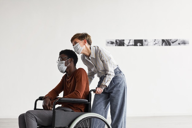 사진 휠체어를 사용하고 마스크를 쓰고 그를 돕는 젊은 여성과 함께 현대 미술관에서 그림을보고있는 아프리카 계 미국인 남자의 최소한의 초상화,