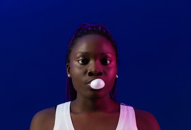 Minimal portrait of young African-American woman blowing bubblegum bubble and looking at camera while standing against purple background in studio