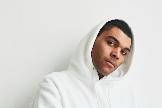 Minimal portrait of young latin american man wearing white\
hoodie and looking at camera against white background, copy\
space