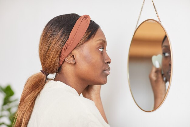 Ritratto minimo con vista laterale di una bella donna afroamericana che si gode la routine di cura della pelle a casa e guarda lo specchio, copia spazio