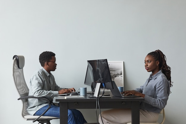 Ritratto con vista laterale minima di due giovani afro-americani che utilizzano computer e scrivono codice mentre lavorano nell'ufficio di sviluppo software, spazio di copia