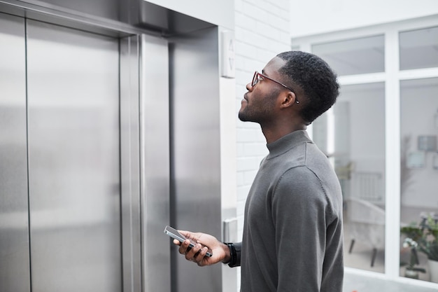 Minimal side view of young black businessman waiting for\
elevator in office building and holding pho