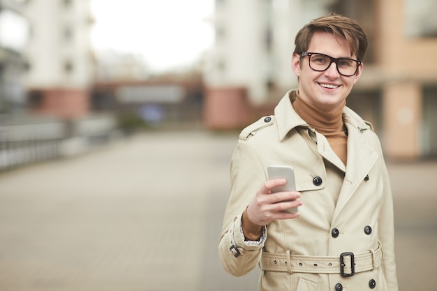 Minimal waist up portrait of modern young businessman wearing trenchcoat and holding smartphone looking at camera while standing outdoors in urban city setting, copy space