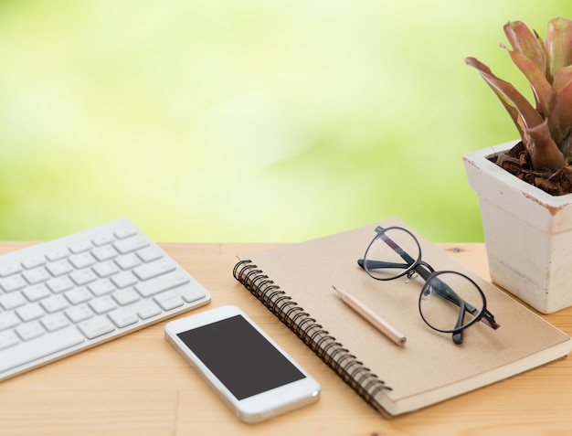 Minimal workspace with keyboard, smartphone, eyeglasses, notebook and pencil on wood table