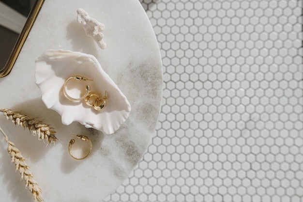 Minimale modesamenstelling met gouden oorbellen in zeeschelp op marmeren tafel met spiegel en tarwestelen