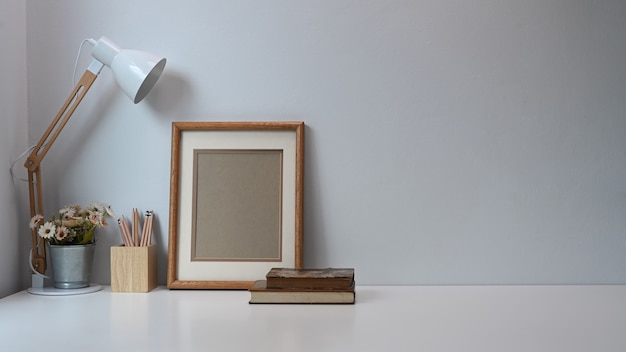 Minimale werkruimte met frame, potloden, koffiekopje, lamp en oud boek op witte tafel.