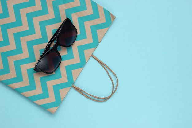 Minimalism shopping concept. Paper shopping bag and sunglasses on blue background.