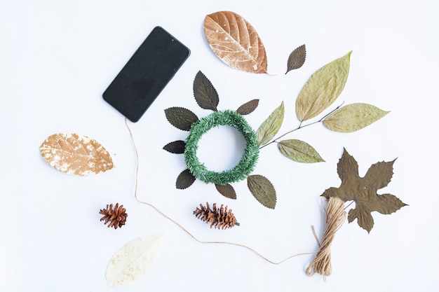 Минималистская осенняя концепция. сушеные листья, сосновые цветы, венок кранс, пряжа из мешковины, смартфон, изолированные на фоне белой бумаги