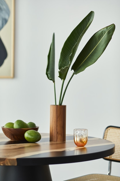 Minimalist composition on the design wooden table with fruits,\
tropical leaf in vase, abstract painitngs and stylish chair. modern\
dining room.