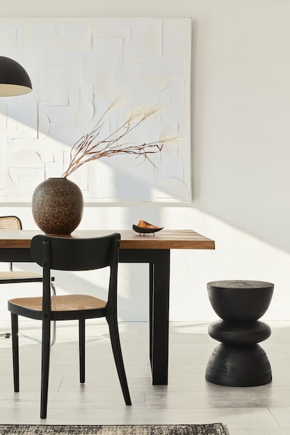 Minimalist composition of dining room interior with wooden table, design chairs, dried flowers in a vase, black pendant lamp, art paintings on the wall and elegant personal accessories.