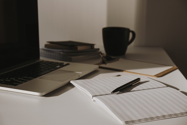 Minimalist home office desk workspace. Laptop on table with coffee cup, paper sheet, stationery against white wall