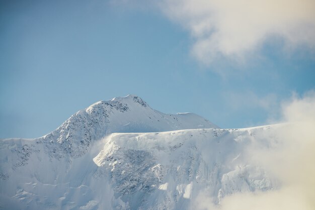 写真 日差しの中で厚い低い雲の中の雪をかぶった山の壁のミニマリストビュー。青い空の濃い雲の中に白い雪のピークがある風光明媚な明るい山の風景。雪に覆われた頂上の素晴らしい景色。