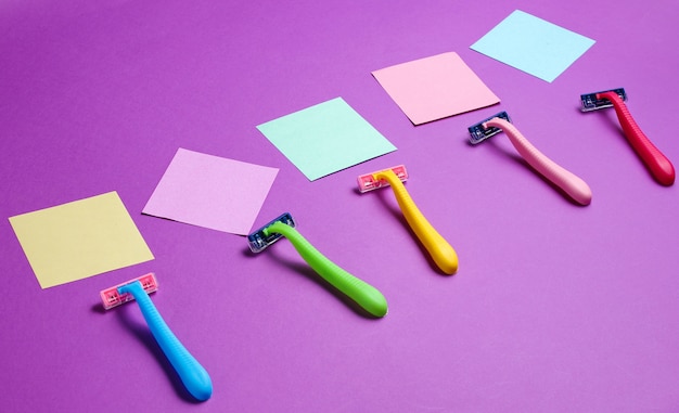 Minimalistic beauty concept. Epilation. Plastic colored razors and memo sheets of paper on purple.