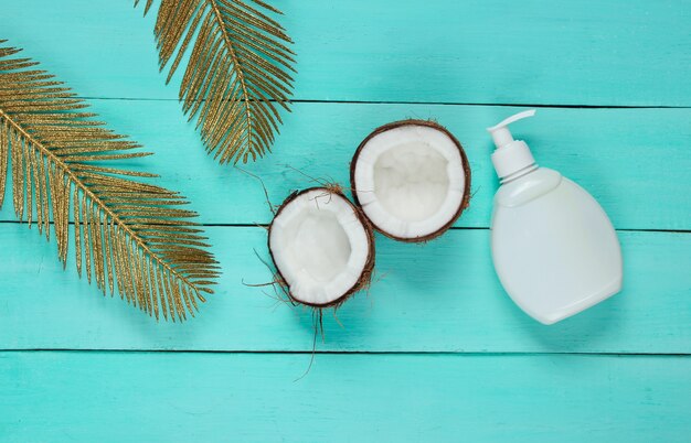 Minimalistic beauty still life. Two halves of chopped coconut and white bottle of cream with golden palm leaves on blue wooden background. Creative fashion concept.