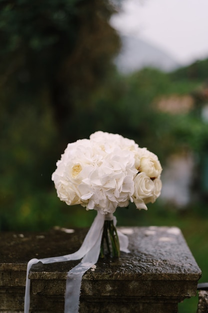 写真 濡れた石の手すりに白いリボンと白いバラとホルテンスのミニマルなブライダルブーケ