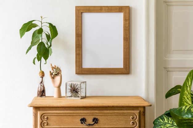 Minimalistic compositon with wooden vintage commode, brown photo frame, avocado plant, plants and elegant personal accessories. Stylish retro living room. White walls..