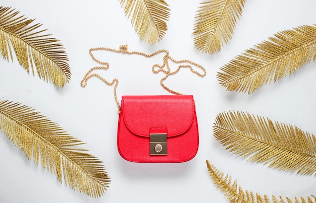 Minimalistic fashion still life. Leather red bag among decorative golden palm leaves on white background. Top view