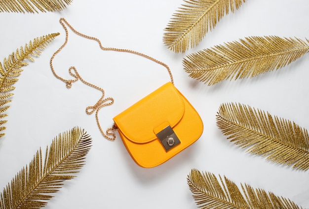 Minimalistic fashion still life. Leather yellow bag among decorative golden palm leaves on a white background. Top view