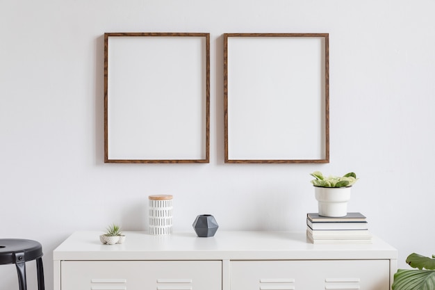 Minimalistic home decor of interior with two brown wooden photo frames on the white shelf with books, beautiful plant in stylish pot and home accessories. White wall.