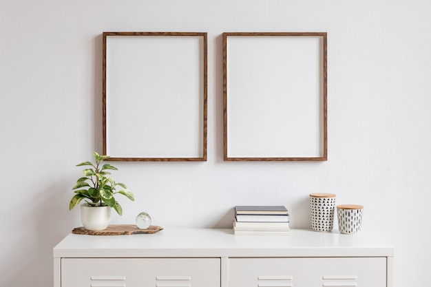 Minimalistic home decor of interior with two brown wooden photo frames on the white shelf with books, beautiful plant in stylish pot and home accessories. White wall.