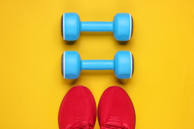 Minimalistic sport still life. sports outfit. red sports shoes\
for training and blue plastic dumbbell on yellow background.