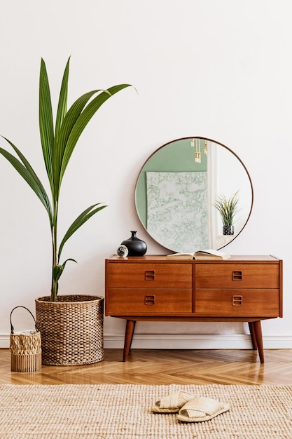 Minimalistic and stylish composition of living room with wooden commode, mirror, chair , lamp, tropical plant in rattan basket and elegant personal accessories. Interior design, Home decor. template.