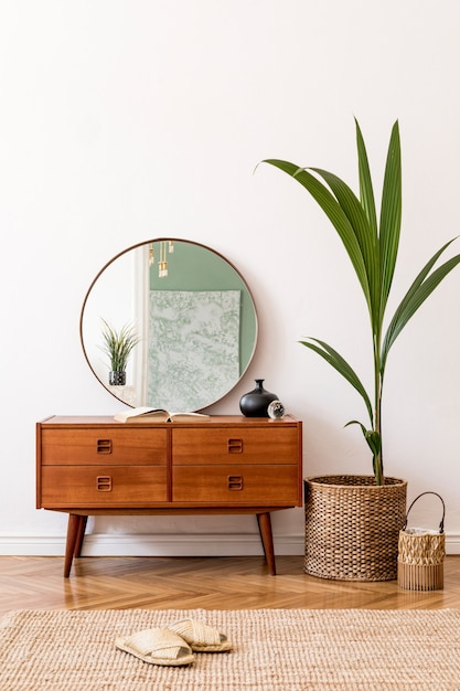 Minimalistic and stylish composition of living room with wooden commode, mirror, tropicla plant in rattan basket and elegant personalaccessories. Interior design, Home decor. White walls.