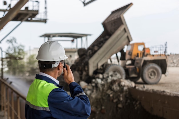 Mining engineer in yellow-blue uniform and helmet supervises unloading dumpers