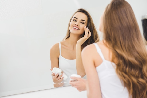 Riflessione speculare di bella donna che applica crema per il viso