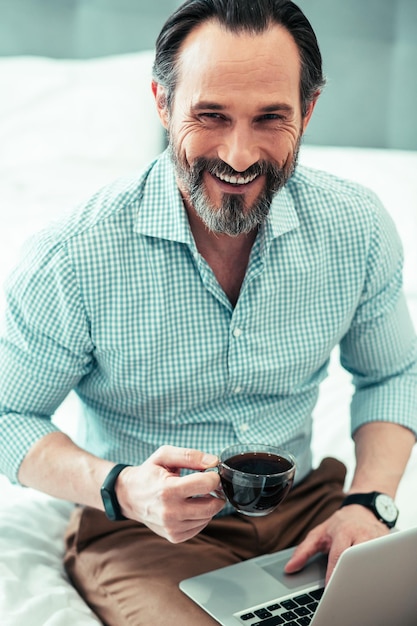 Mirthful bearded man holding a cup of coffee and laughing while sitting with a laptop