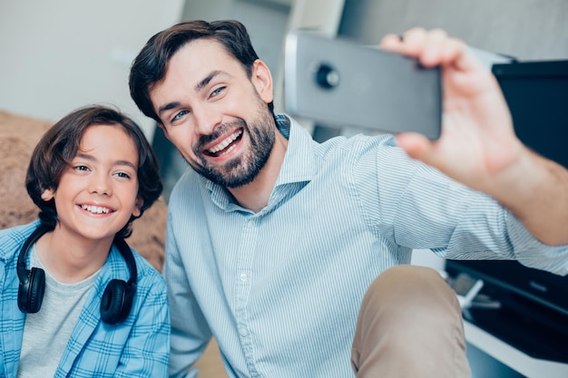 Mirthful boy smiling while his positive parent holding a smartphone and taking nice photos with him