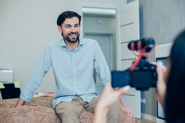 Mirthful handsome man sitting on a bed at home and smiling to the camera with a woman recording him